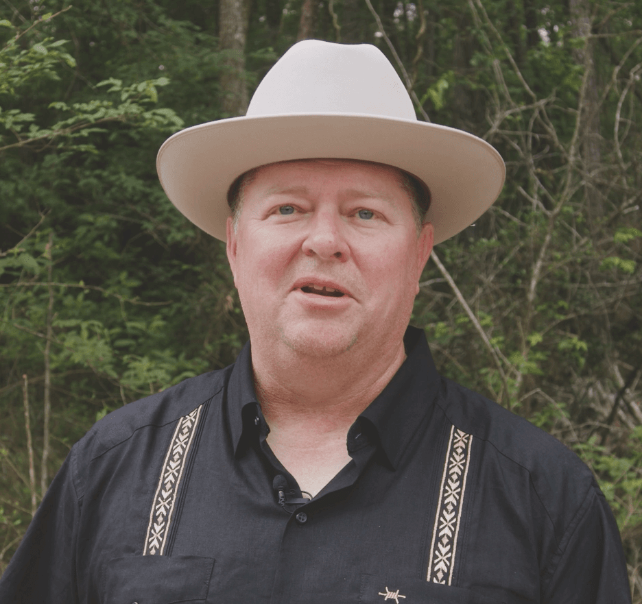 A middle-aged man wearing a light-colored cowboy hat and a black shirt stands outdoors in front of trees and greenery at TX at Two Step Farm in Montgomery County, TX.