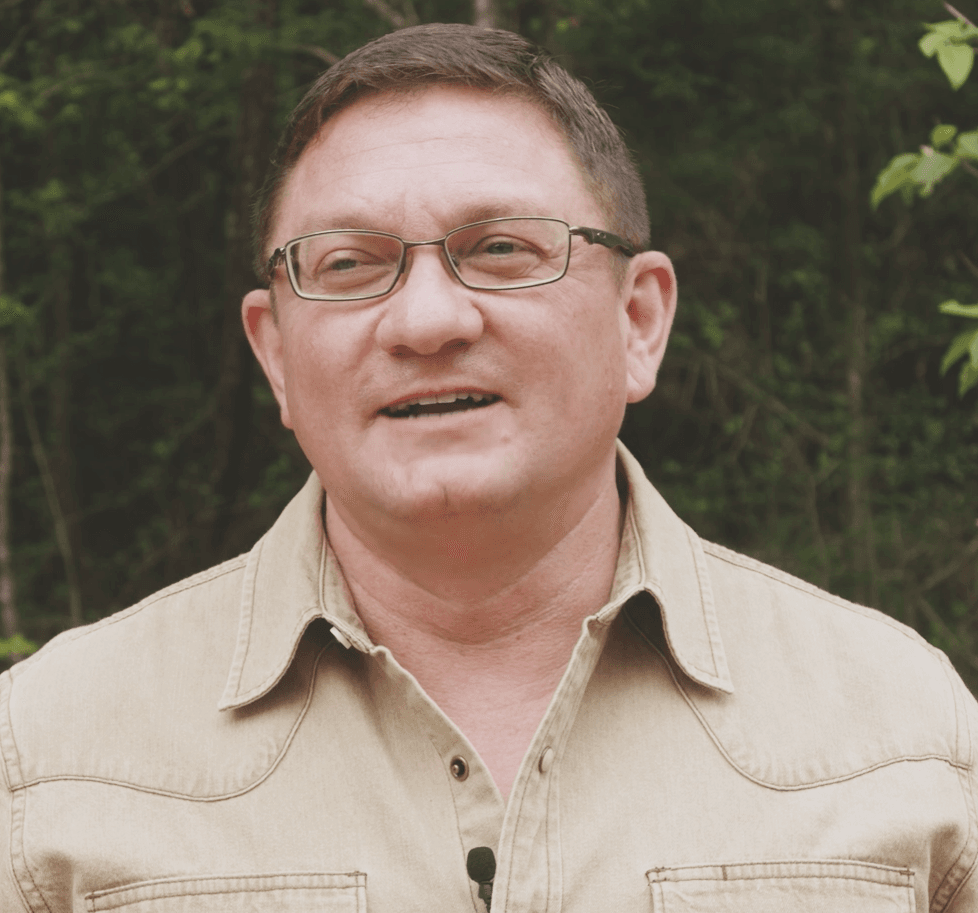 A man wearing glasses and a beige shirt stands outdoors in front of green foliage at TX at Two Step Farm in Montgomery County TX, speaking with a small microphone clipped to his shirt.
