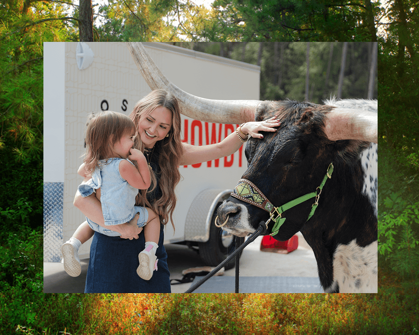 A woman holding a smiling child pets a large longhorn cow wearing a patterned halter at The Grazery restaurant at Two Step Farm. A trailer, trees, and the charm of TX’s new homes in Montgomery County set the scene.