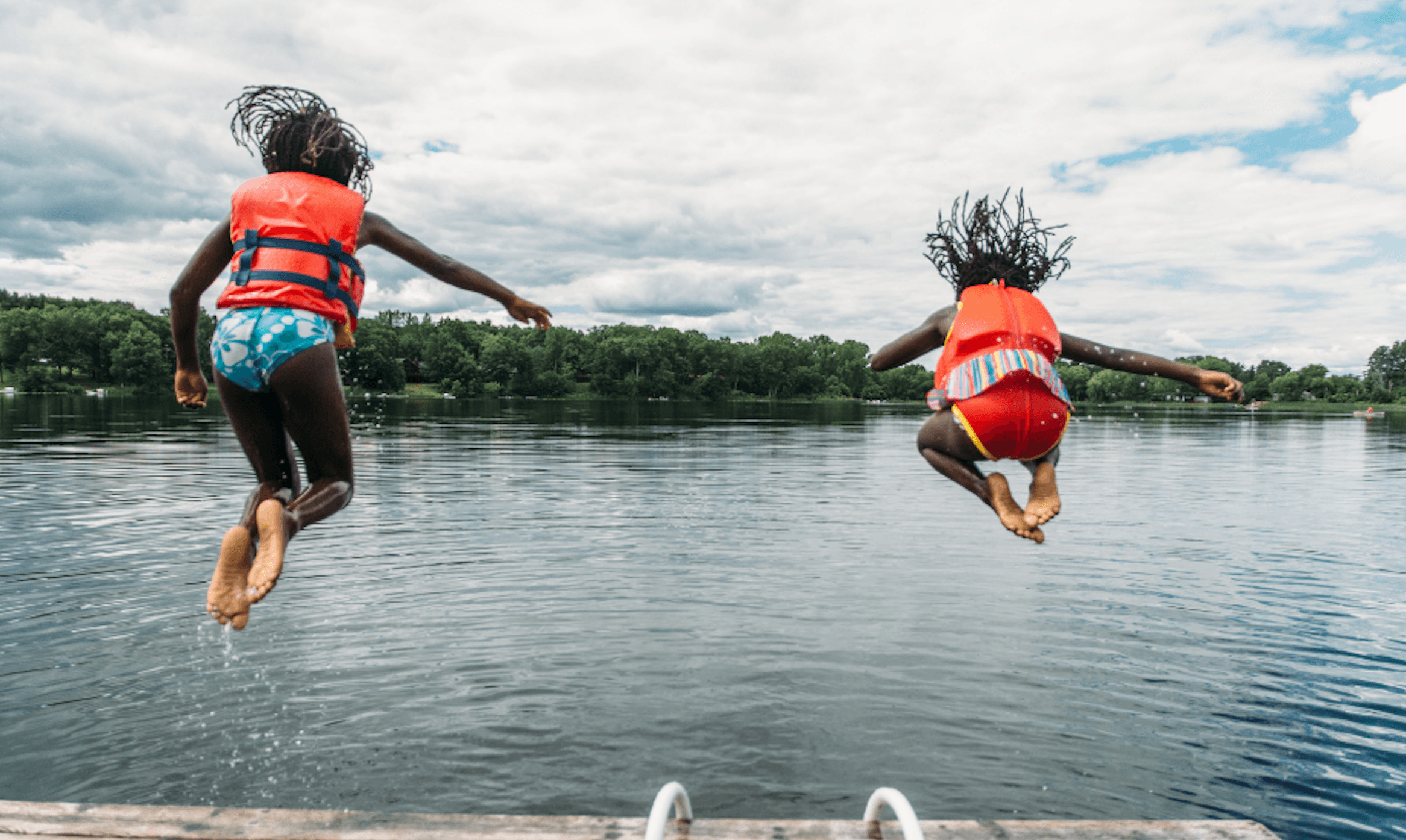 Two children wearing life jackets jump off a dock into a lake near the new homes at Two Step Farm, with trees and a cloudy sky in the background.