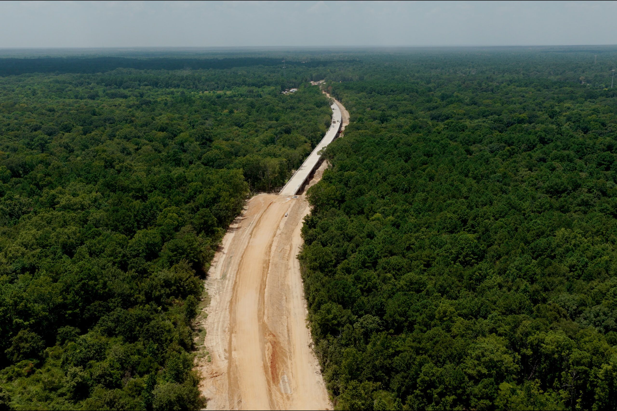 Aerial view of a wide dirt road or construction site cutting through dense green forest, with a partially built structure along its length.