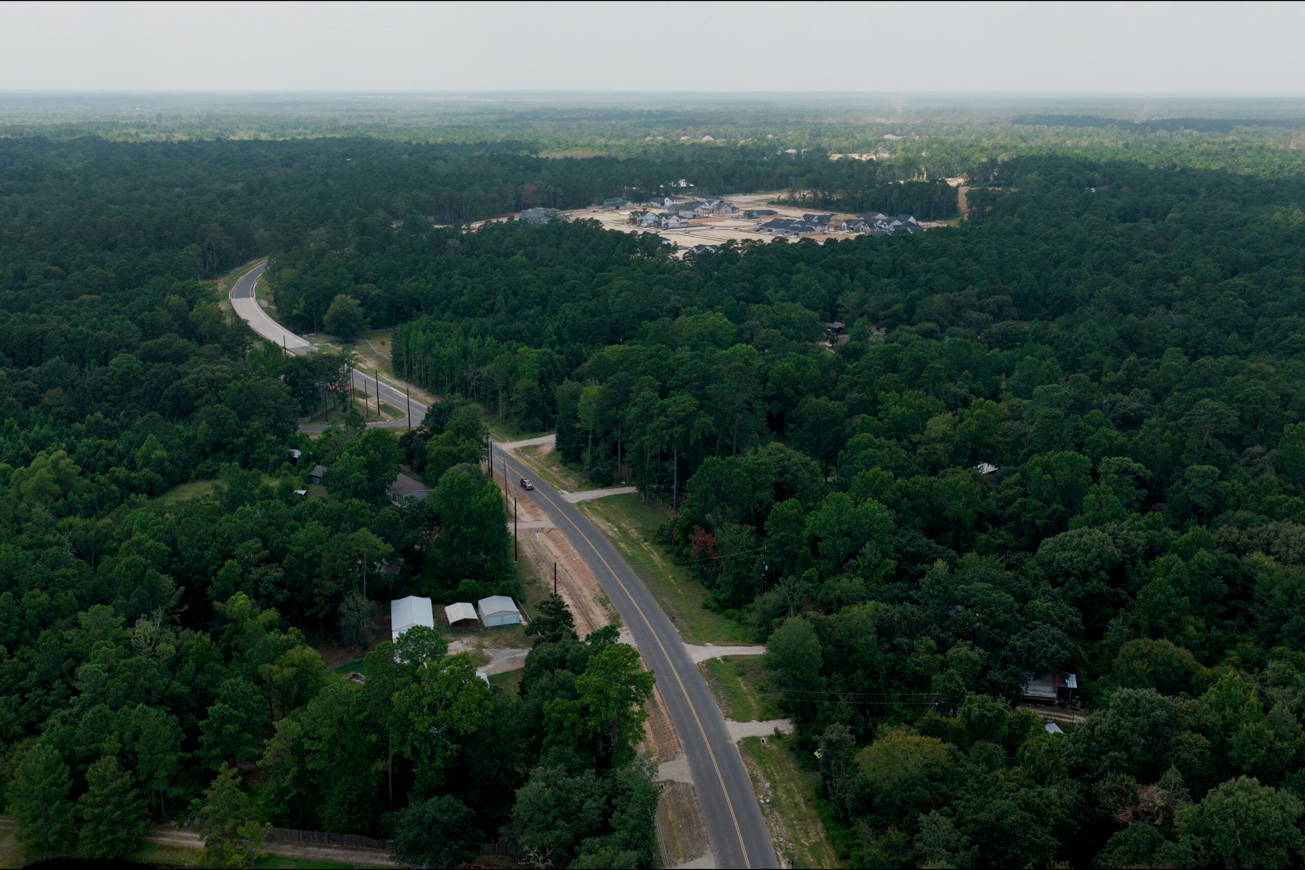 A winding road runs through a dense, green forest with scattered houses, leading to a construction site in the distance under a hazy sky.