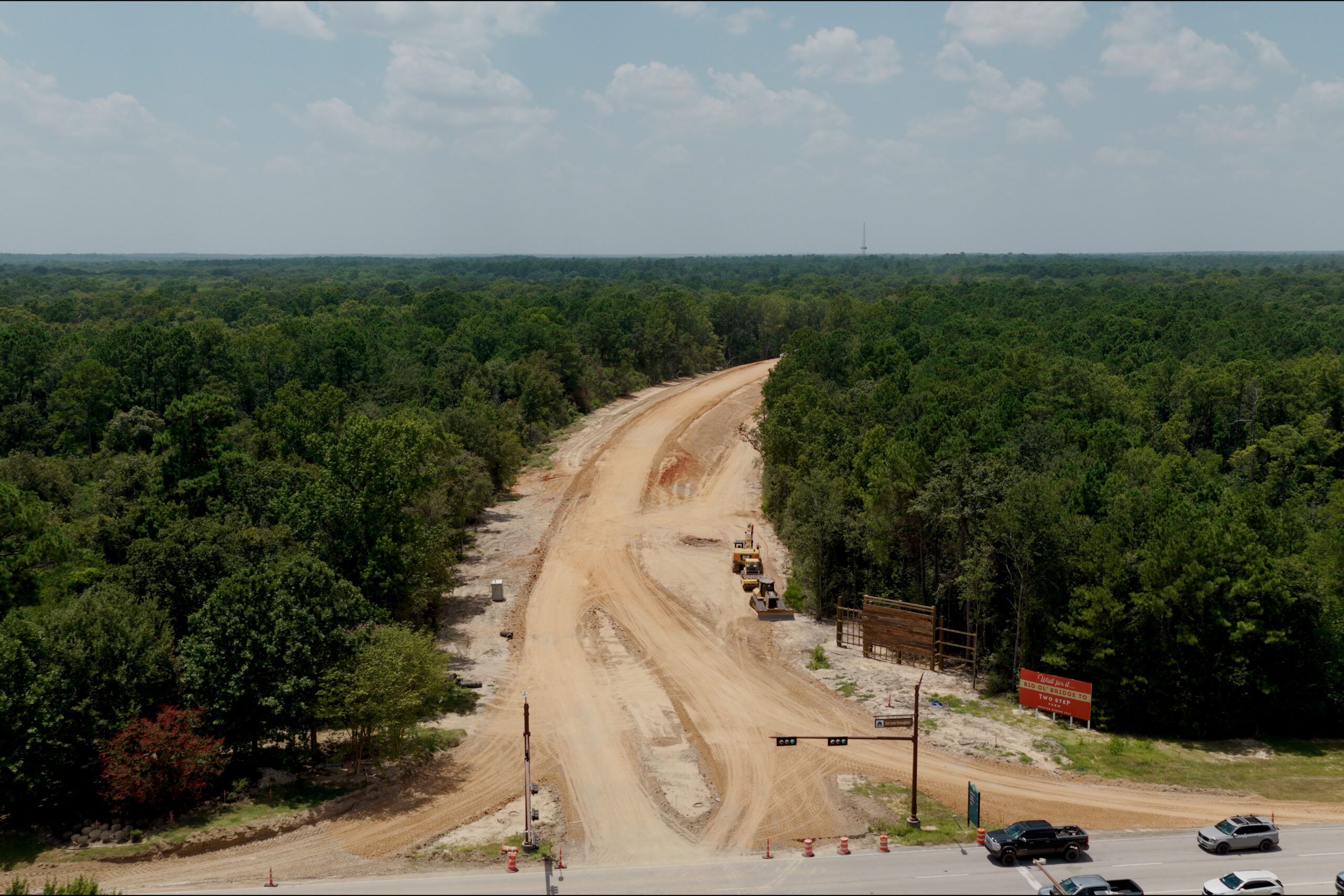 Aerial view of a partially constructed road through a forested area, with construction vehicles and equipment on the site. Several cars are stopped at an intersection in the foreground.