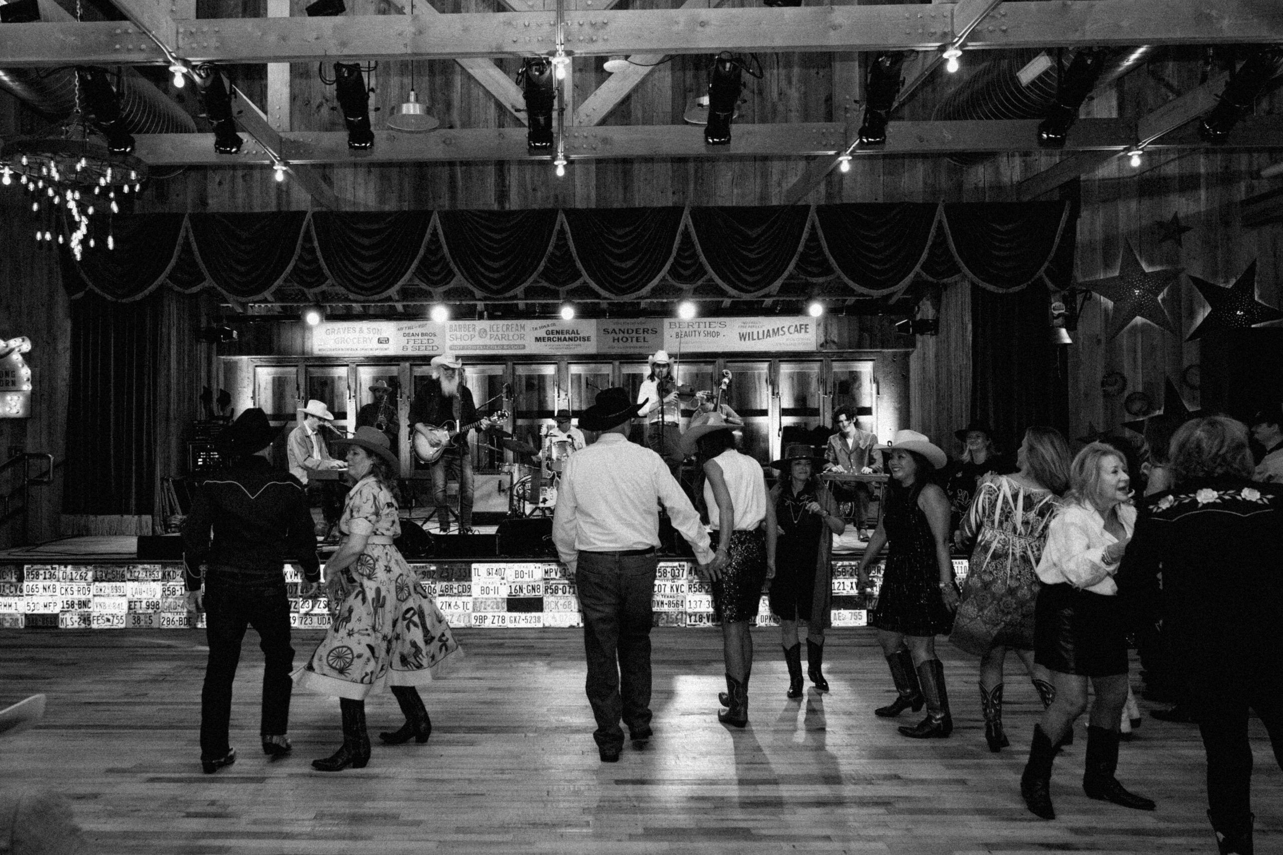 People dressed in Western attire dance in pairs on a wooden floor while a band performs on stage at The Grazery restaurant at Two Step Farm, creating the perfect country-themed night out.