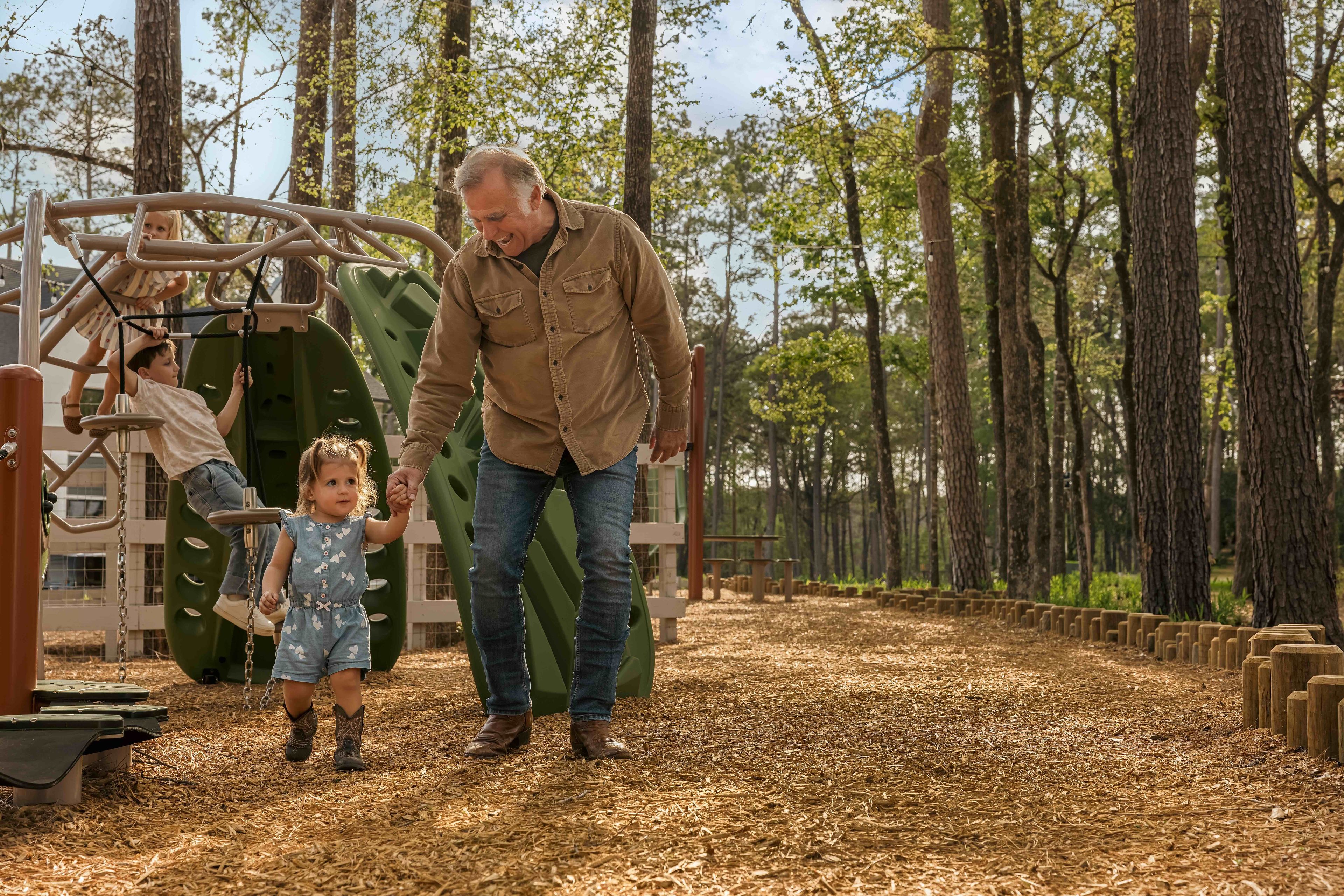An older man holds a toddler's hand while walking on a playground path near The Grazery restaurant at Two Step Farm; another child plays on climbing equipment in the background among tall trees.