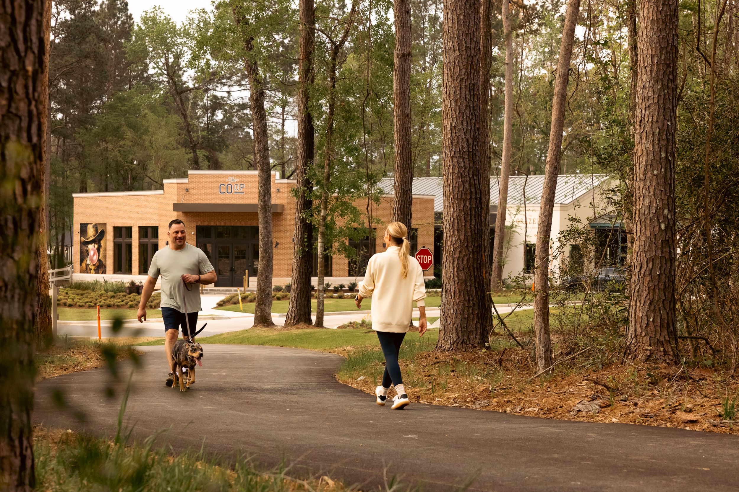 A man jogs with a dog and a woman walks on a paved path through trees near a modern brick building with a sign reading "COOP," capturing the vibrant community spirit near new homes in Montgomery County, TX.