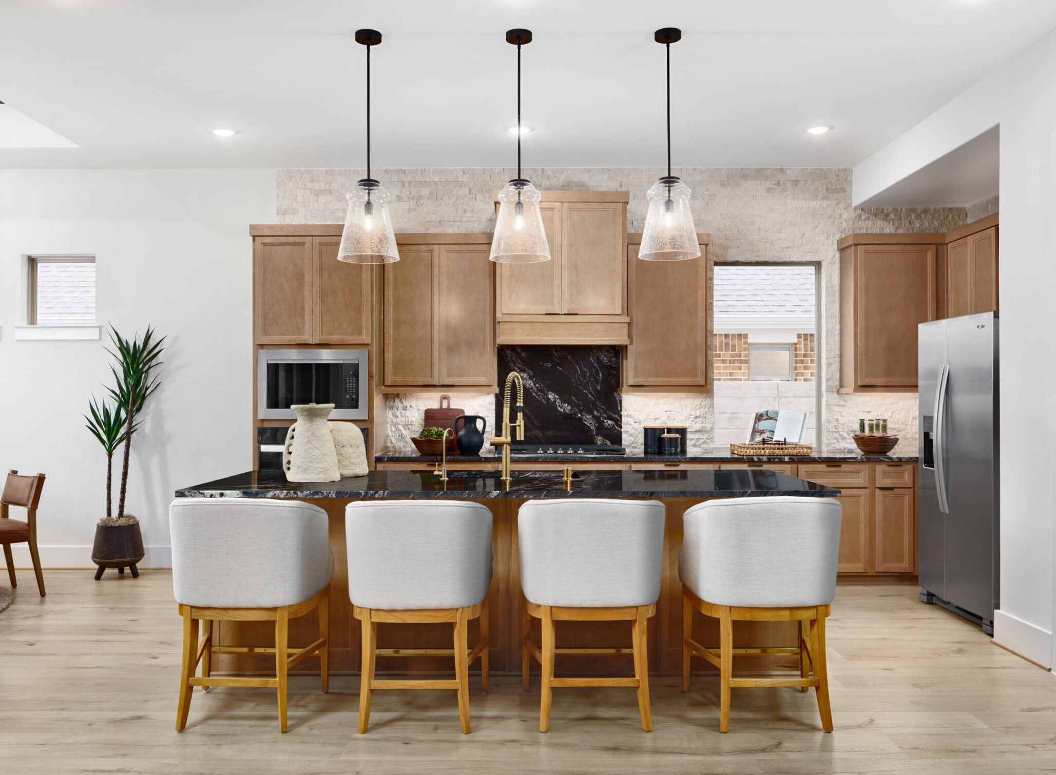 Modern kitchen with light wood cabinets, a black island with four white barstools, pendant lights, stainless steel appliances, and a plant by the dining area.
