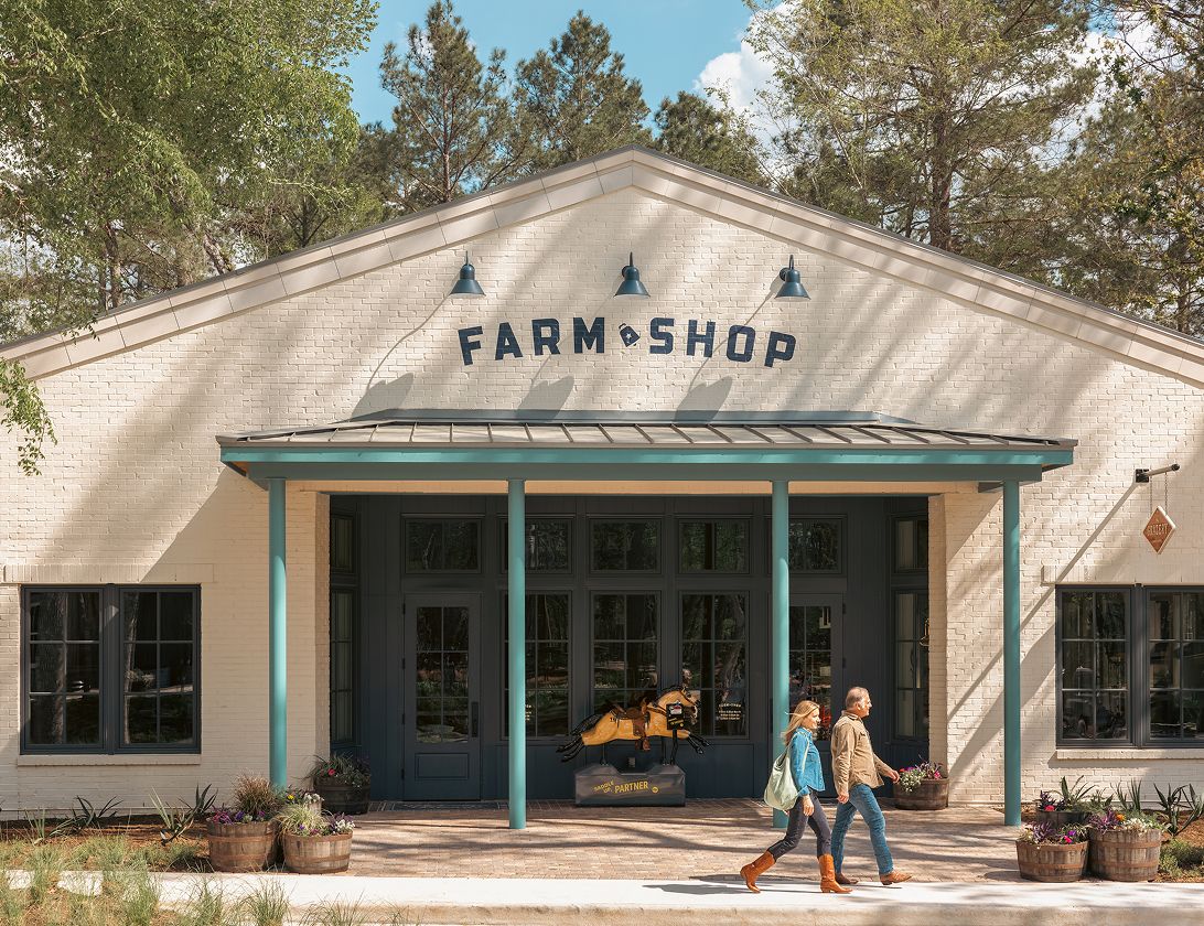 Two people walk past a white building with large windows and a sign above the entrance that reads “Farm Shop.” Trees and plants surround the area.